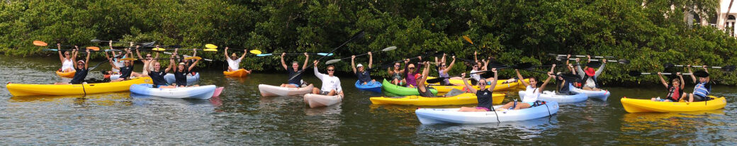 A group photo of around 26 volunteers both male and female. They are all floating in the river on kayaks. All kayaks are different colors. They are all holding their kayak paddles above their heads and smiling. They are in front of a large stretch of mangroves.